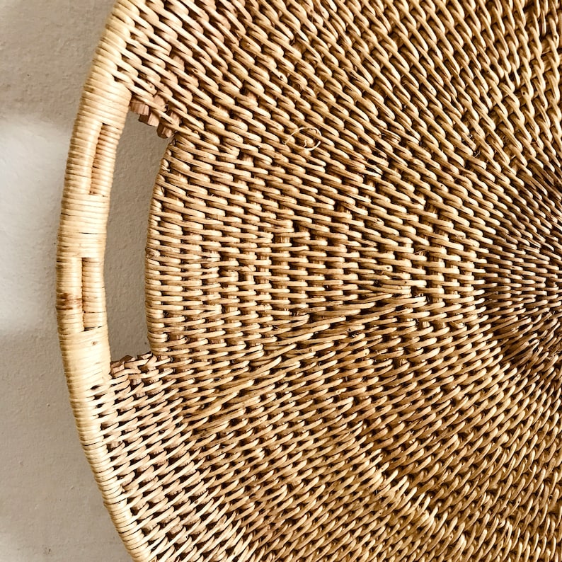 May include: A round, woven wicker tray with a handle. The tray is made of natural-colored wicker and has a tightly woven pattern. The handle is a curved shape, and the tray is likely used for serving or decoration.