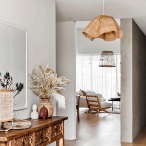 May include: A wooden console table with three drawers, a vase with dried flowers, and a woven lampshade hanging from the ceiling. The table is in a hallway with a white wall and a doorway leading to another room.