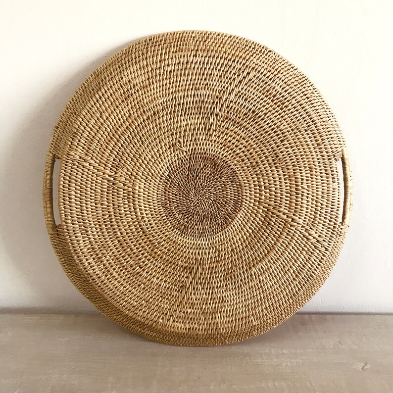 May include: A round, woven tray with two handles. The tray is made of natural-colored rattan and has a circular design. The handles are integrated into the tray's design, providing a functional and decorative element. The tray is suitable for serving food or as a decorative piece.