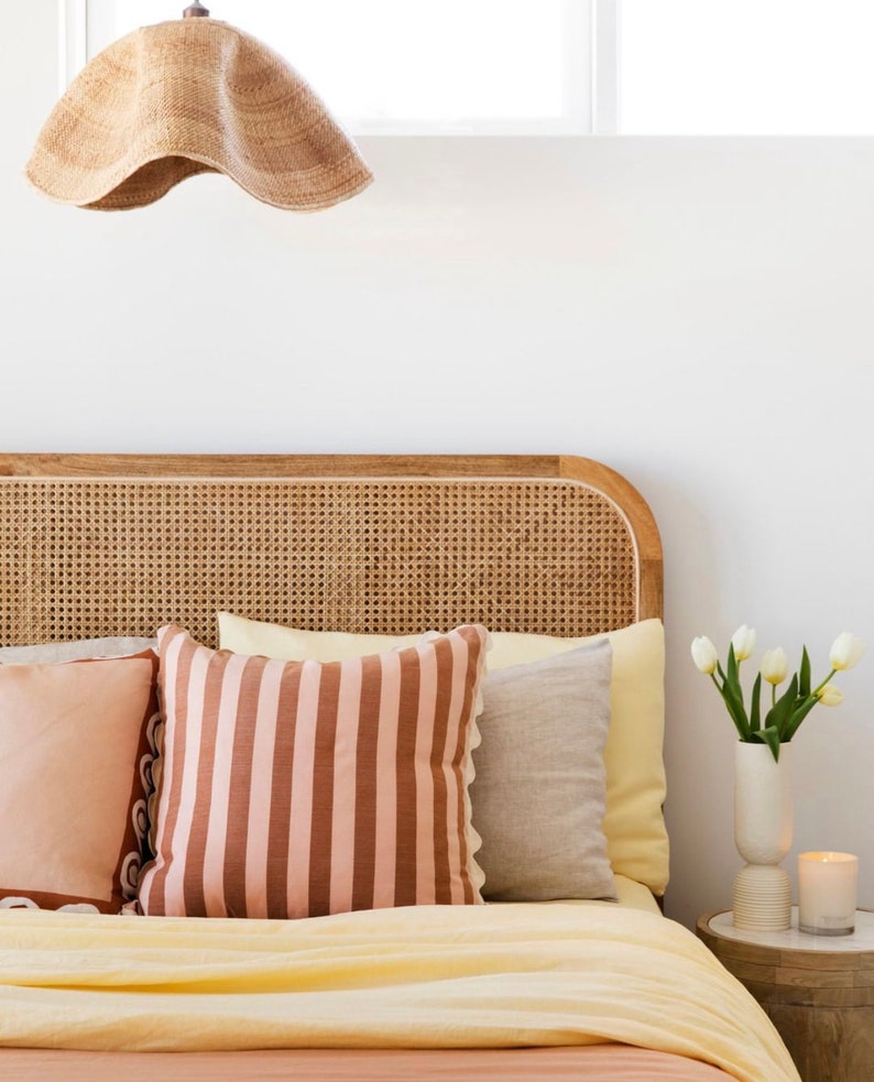May include: A bedroom with a bed featuring a woven headboard, a yellow and pink striped pillow, and a white vase with white tulips on a wooden nightstand.
