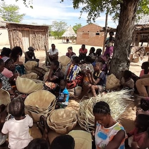May include: A community of people, including children, are gathered outside, possibly in a village. They are crafting large, light-coloured woven baskets. The scene suggests traditional craftwork being undertaken by the community.