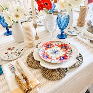 Può includere: Una tavola splendidamente apparecchiata con piatti floreali bianchi e blu, bicchieri blu e vasi bianchi con fiori rossi e bianchi. Tovagliette intrecciate, candele e posate con manici in bambù completano l'elegante scena.