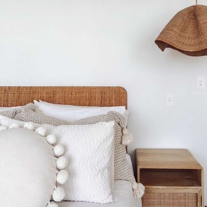 May include: A white bed with a woven headboard and a white, beige, and brown patterned throw pillow. The bed has a white duvet cover and a white pillow with pom-poms. A wooden nightstand with a woven drawer is next to the bed.