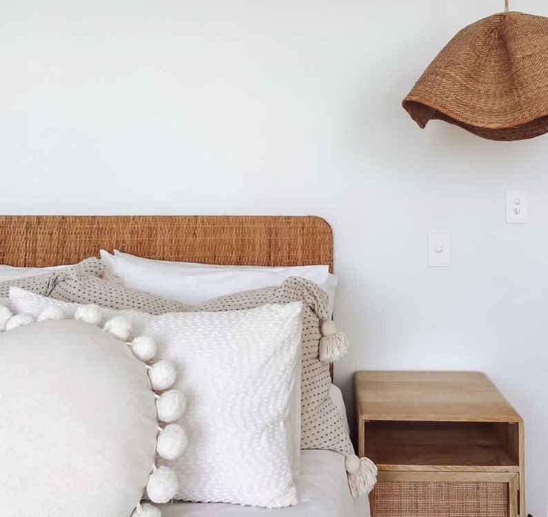 May include: A white bed with a woven headboard and a white, beige, and brown patterned throw pillow. The bed has a white duvet cover and a white pillow with pom-poms. A wooden nightstand with a woven drawer is next to the bed.