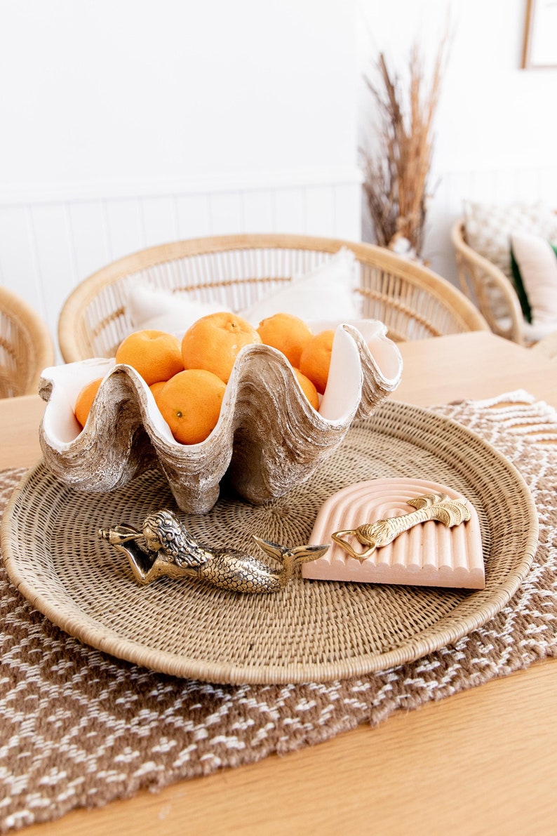 May include: A decorative arrangement featuring a large, clam-shaped bowl filled with oranges. A woven tray holds a mermaid bottle opener and a pink, arched coaster with a gold accent. The scene is set on a wooden table.