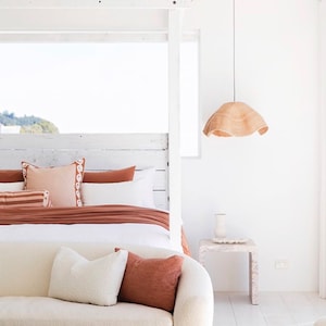 May include: A white couch with a white throw pillow and a rust-colored throw pillow sits in front of a white bed with a rust-colored duvet and white pillows. A white nightstand with a vase sits next to the bed. A woven pendant light hangs from the ceiling.