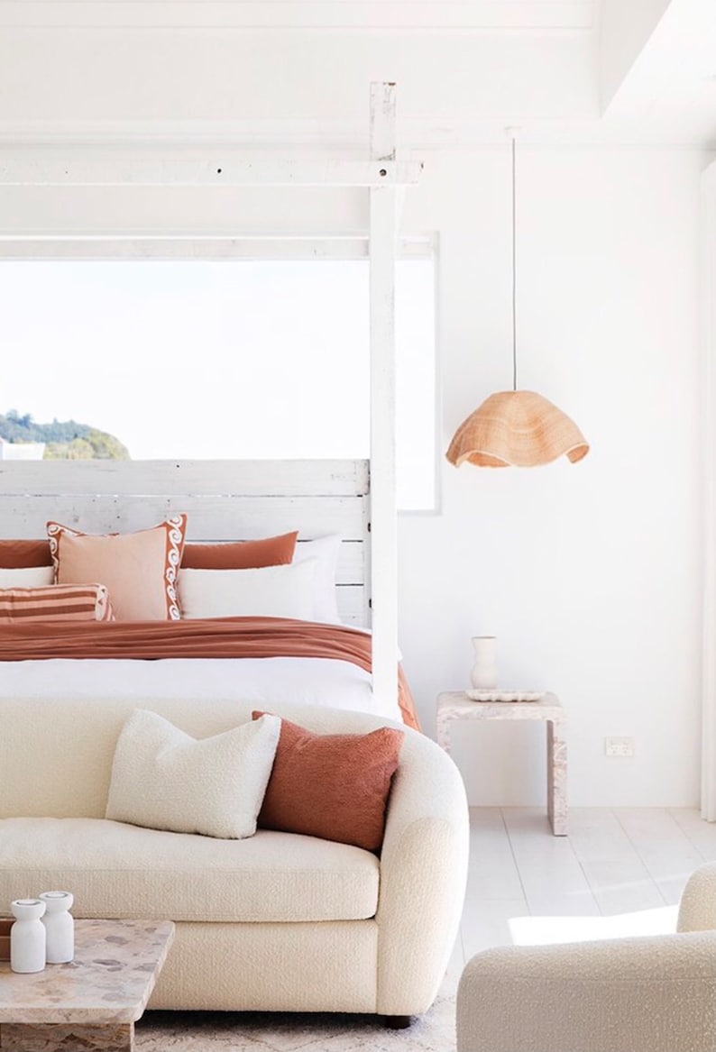 May include: A white couch with a white throw pillow and a rust-colored throw pillow sits in front of a white bed with a rust-colored duvet and white pillows. A white nightstand with a vase sits next to the bed. A woven pendant light hangs from the ceiling.