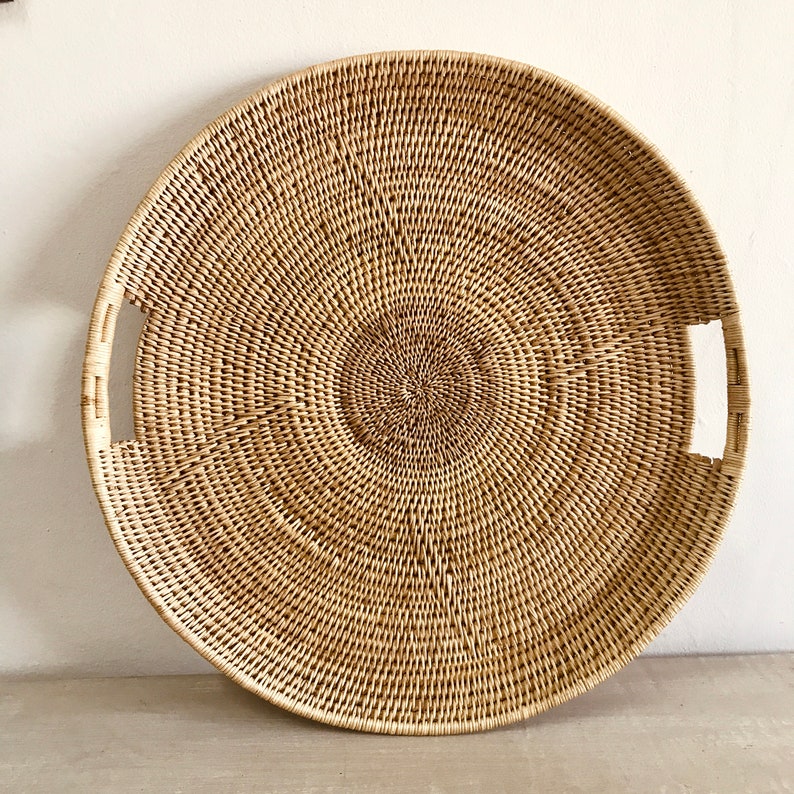 May include: A large, round woven tray with a natural, light brown color. The tray features a circular design with two rectangular handles on opposite sides. The woven pattern creates a textured surface, suitable for serving or display.