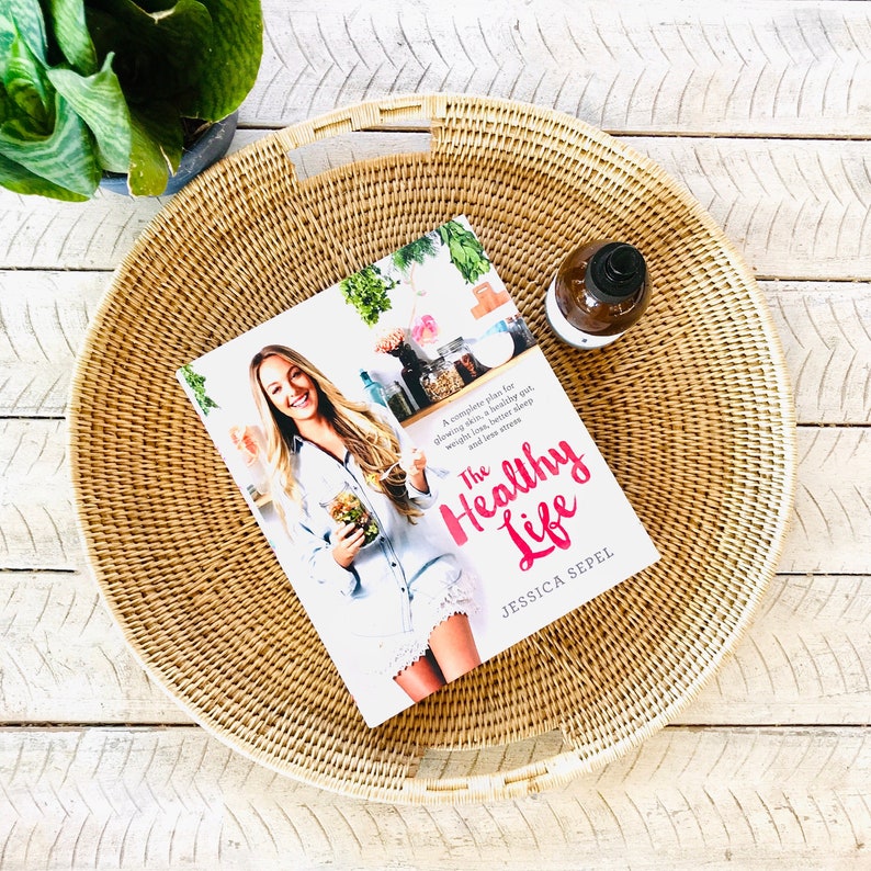 May include: A round, woven wicker tray with handles holds a copy of "The Healthy Life" by Jessica Sepel, a book about health and wellness. A small brown bottle with a black top sits on the tray. A green plant is in the background.