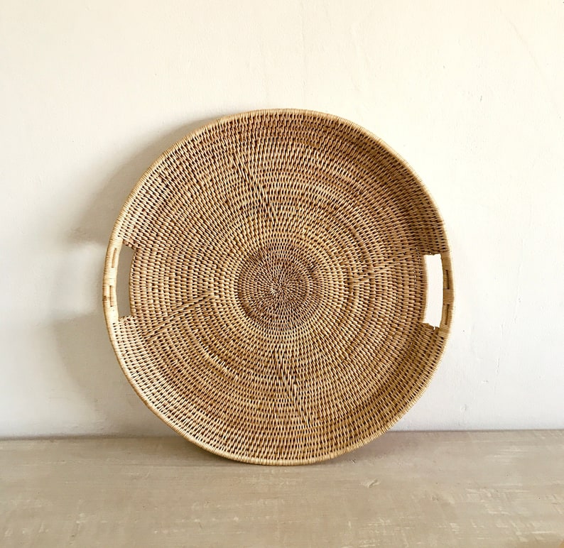 May include: A round, woven rattan tray with a natural, light brown color. The tray features a central circular design and two rectangular handles. The woven pattern creates a textured surface. This decorative tray is suitable for serving or display.