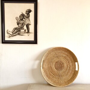 May include: A framed black and white pencil drawing of two children, one combing the other's hair. Below, a large, round, woven basket with handles rests on a light-colored surface against a white wall.