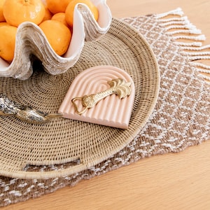 May include: A still-life image featuring a decorative arrangement. A white clam shell bowl holds several oranges. A pink arched bottle opener with a gold palm tree design rests on a woven tray. The tray sits on a patterned placemat.