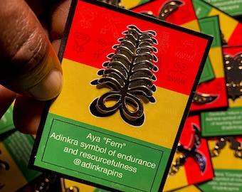 May include: A gold-colored pin with a fern design, representing the Adinkra symbol "Aya" which symbolizes endurance and resourcefulness. The pin is on a card with the text "Aya "Fern" Adinkra symbol of endurance and resourcefulness @adinkrapins".
