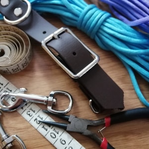 May include: A dark brown leather strap with a silver metal buckle, two silver metal swivel snap hooks, a white measuring tape showing inches, and two coils of bright blue and purple paracord, accompanied by a pair of black needle-nose pliers.