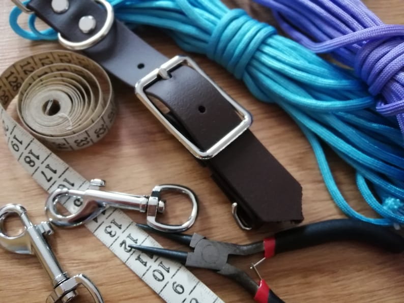 May include: A dark brown leather strap with a silver metal buckle, two silver metal swivel snap hooks, a white measuring tape showing inches, and two coils of bright blue and purple paracord, accompanied by a pair of black needle-nose pliers.