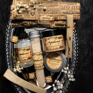 Puede incluir: Una bandeja de plata con una variedad de productos de botica, que incluyen una botella de aceite corporal, un frasco de exfoliante corporal, un tubo de sales de baño, un frasco de exfoliante labial y un palo de incienso. Los productos están todos etiquetados con la palabra "Apothecary" y tienen una estética vintage.