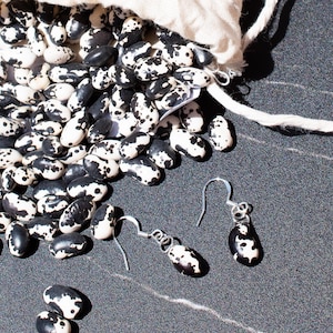 May include: A white cloth bag spills out black and white speckled beans. Two pairs of earrings dangle below, each featuring a single bean as a pendant.