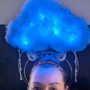 May include: A whimsical headpiece featuring a fluffy, glowing blue cloud. The cloud is adorned with small lights, creating a dreamy effect. The headpiece includes a delicate silver headband with dangling crystal accents, perfect for a costume or themed event.