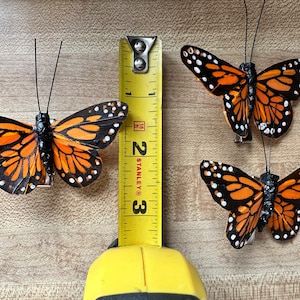 Peut inclure: Trois ornements papillon monarque avec des ailes orange, noires et blanches et de fines antennes noires. Un m&egrave;tre ruban jaune Stanley est plac&eacute; entre les papillons, montrant que le plus grand papillon a une envergure d'environ 7,6 cm.