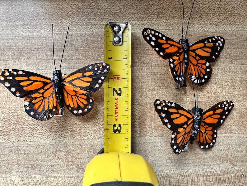 Peut inclure: Trois ornements papillon monarque avec des ailes orange, noires et blanches et de fines antennes noires. Un m&egrave;tre ruban jaune Stanley est plac&eacute; entre les papillons, montrant que le plus grand papillon a une envergure d'environ 7,6 cm.