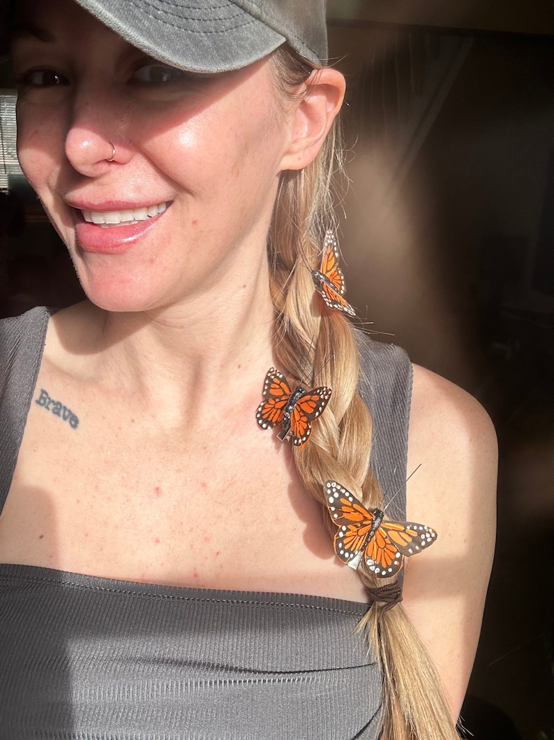 May include: Three orange and black butterfly hair clips in a blonde braid. The braid is held together with a brown hair tie. The word 'Brave' is tattooed on the wearer's left shoulder.
