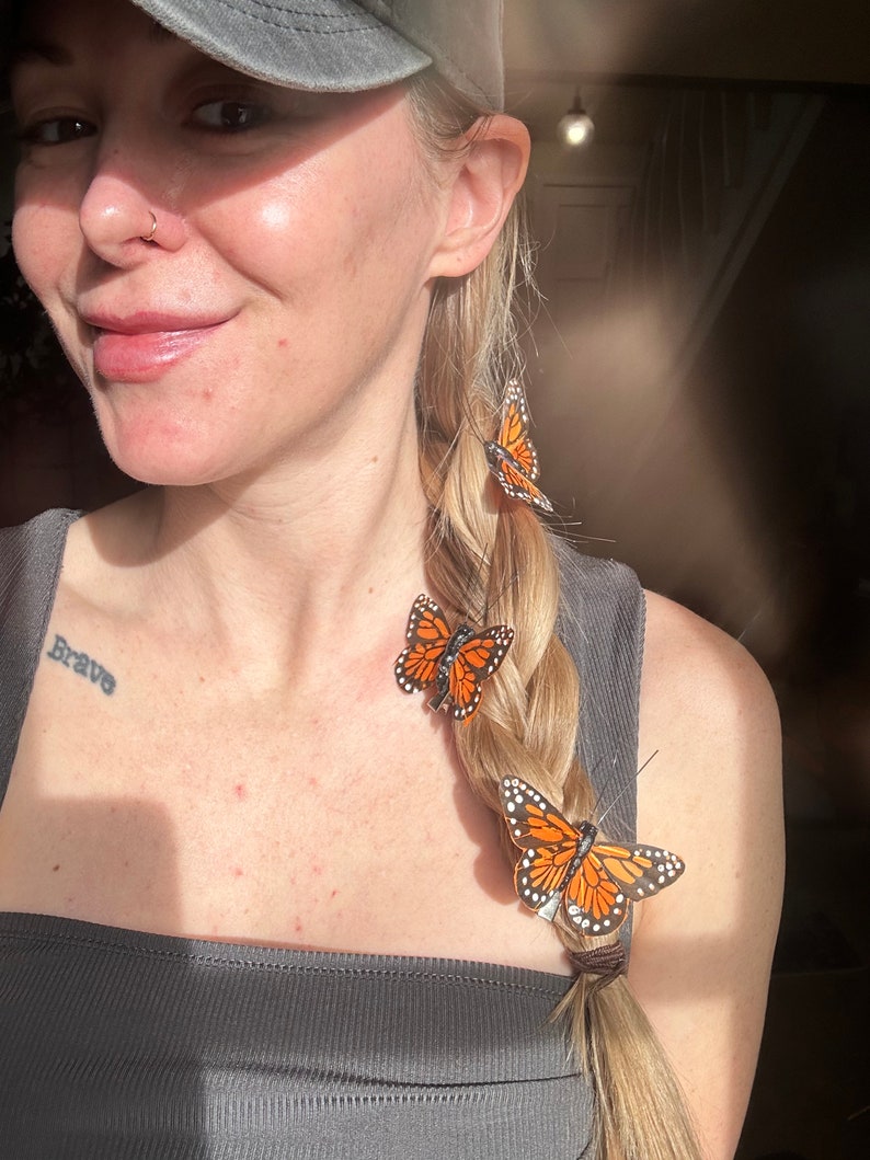 Peut inclure: Trois pinces &agrave; cheveux en forme de papillon monarque avec des ailes orange, noires et blanches fix&eacute;es sur une tresse blonde. Les papillons pr&eacute;sentent des motifs d'ailes r&eacute;alistes et de fines antennes, fix&eacute;s aux cheveux par des pinces m&eacute;talliques.