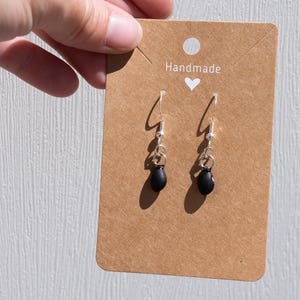 May include: Handmade black dangle earrings with silver hooks. The earrings are displayed on a brown card with the word "Handmade" and a heart.