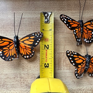 Peut inclure: Trois ornements papillon monarque avec des ailes orange, noires et blanches, chacun dot&eacute; de fines antennes noires et d'une pince m&eacute;tallique sur le dessous. Un m&egrave;tre ruban jaune Stanley est plac&eacute; verticalement, indiquant une longueur de 7,6 cm.