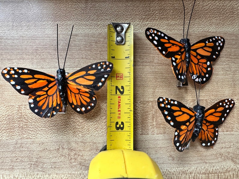 Peut inclure: Trois ornements papillon monarque avec des ailes orange, noires et blanches, chacun dot&eacute; de fines antennes noires et d'une pince m&eacute;tallique sur le dessous. Un m&egrave;tre ruban jaune Stanley est plac&eacute; verticalement, indiquant une longueur de 7,6 cm.