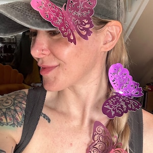 Peut inclure: Une femme portant un anneau au nez porte une casquette de baseball noire avec un clip à cheveux papillon rose sur le côté. Elle a une tresse dans les cheveux avec deux autres clips à cheveux papillon rose. Les papillons ont un design délicat et complexe.