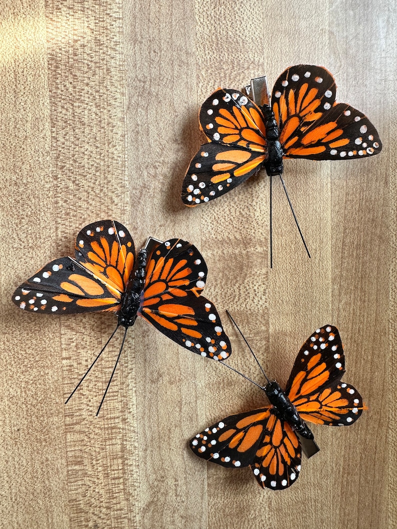 Peut inclure: Trois pinces &agrave; cheveux papillon avec des ailes orange et noires orn&eacute;es de points blancs. Chaque pince poss&egrave;de un corps noir et deux fines antennes. Les ailes ont un aspect textur&eacute; et le m&eacute;canisme m&eacute;tallique est visible en dessous.