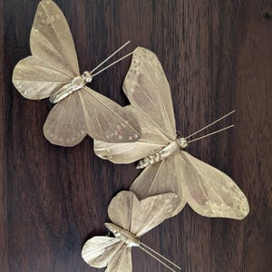 Peut inclure: Trois décorations de papillons dorés avec des détails d'ailes délicats et de fines antennes. Les papillons sont disposés sur une surface en bois brun foncé, créant un contraste de couleurs et de textures. Ces papillons décoratifs sont parfaits pour l'artisanat ou la décoration intérieure.