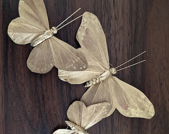 May include: Three gold-colored butterfly decorations with delicate wing details and slender antennae. The butterflies are arranged on a dark brown wooden surface, creating a contrast in color and texture. These decorative butterflies are ideal for crafts or home decor.