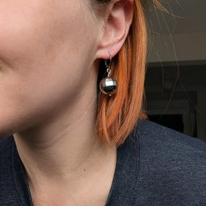 May include: A silver disco ball earring with a lever back closure. The earring is a sphere with multiple reflective facets. The earring is shown on a person's ear, with orange hair in the background.