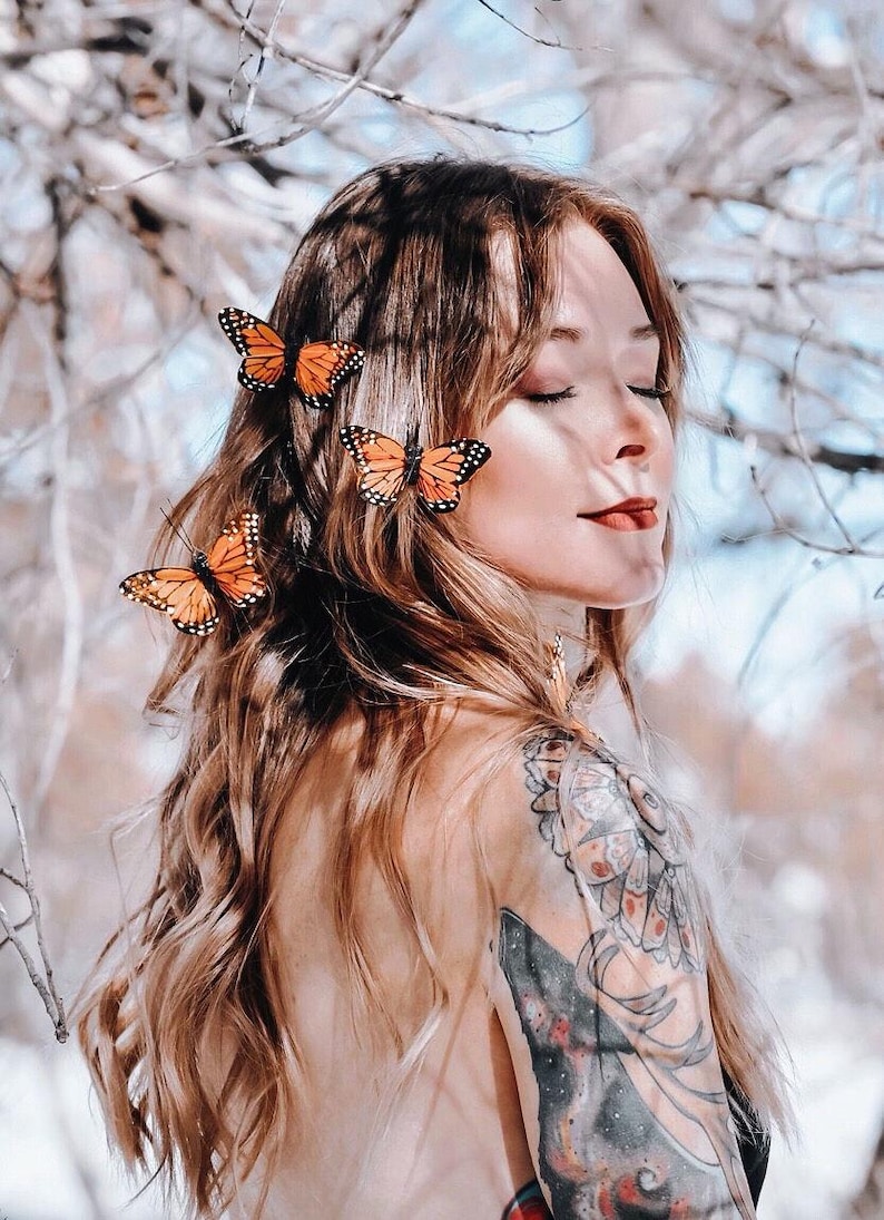 Peut inclure: Trois accessoires de cheveux en forme de papillon monarque orange et noir fix&eacute;s sur de longs cheveux bruns ondul&eacute;s. Les cheveux pr&eacute;sentent un tatouage visible sur l'&eacute;paule avec des motifs floraux et g&eacute;om&eacute;triques complexes en noir, gris et orange.