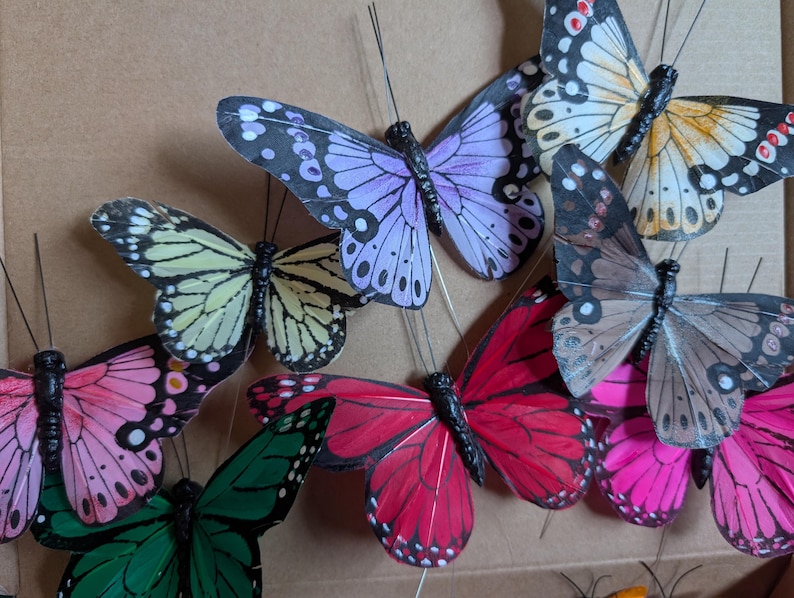 Peut inclure: Une collection de papillons artificiels avec de fines antennes en fil de fer, pr&eacute;sentant diverses couleurs dont le violet clair, le jaune p&acirc;le, le rose vif, le vert profond et des motifs bruns avec des bords d'ailes noirs et des taches blanches.