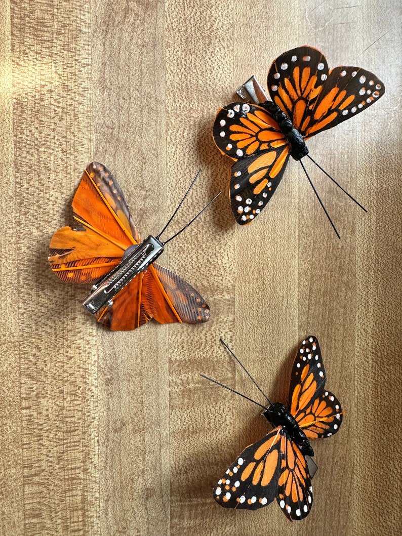 Peut inclure: Trois pinces &agrave; cheveux en forme de papillon avec des ailes orange et noires orn&eacute;es de points blancs. Chaque pince est dot&eacute;e d'une attache m&eacute;tallique de type crocodile fix&eacute;e sous le papillon, incluant deux fines antennes en fil noir.