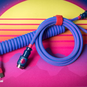Puede incluir: Un cable en espiral azul y rosa con un conector USB en un extremo y un conector redondo en el otro extremo. El cable está en espiral y tiene una correa roja alrededor.