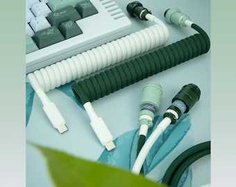 May include: Two coiled keyboard cables, one white and one green, with USB-C connectors. The cables are coiled and have a braided design. The cables are on a light blue surface with a green leaf in the background.