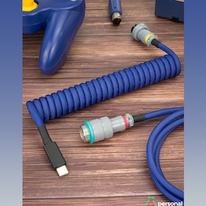 以下が含まれることがあります： 片側にUSB-Cコネクタ、もう片側にPS/2コネクタが付いた青いコイル状のキーボードケーブル。ケーブルはコイル状になっており、赤いリングが付いた灰色のコネクタが付いています。ケーブルは木製の表面にあります。