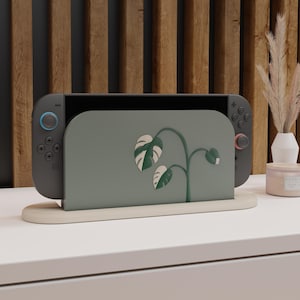May include: A gray Nintendo Switch console with blue and red controllers is displayed in a light wood stand. The console has a green cover with a white and green monstera leaf design. The stand is on a white surface.