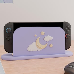 May include: A lavender gaming console dock featuring a raised, three-dimensional design of a pale yellow crescent moon, white clouds, and small yellow stars on the front panel. The dock holds a black gaming console with dark gray controllers.