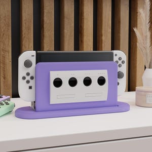 May include: Purple Nintendo Switch dock with a white GameCube controller adapter. The dock has four slots for GameCube controllers and a space for the Switch console. The dock is on a white surface.