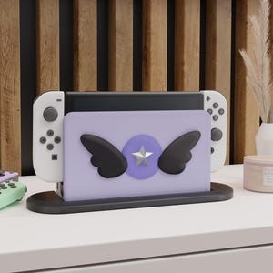 Peut inclure: Une station d'accueil pour console de jeu couleur lavande, ornée d'un cercle violet central avec une étoile argentée et deux ailes noires fixées à l'avant. Le support est posé sur une base noire et contient une console blanche avec des manettes.