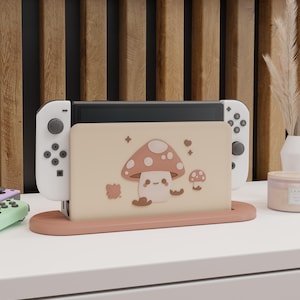 Puede incluir: Una consola Nintendo Switch con mandos blancos está acoplada en un soporte de color rosa pálido. La pantalla de la consola está cubierta por una funda beige con un diseño de hongo de dibujos animados. El soporte está sobre una superficie blanca.
