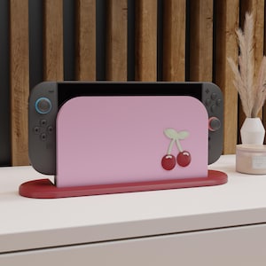 Op de afbeelding: Een Nintendo Switch-console staat op een rode standaard, met een roze hoes met een kersendesign. De console is grijs met blauwe en rode knoppen. De achtergrond omvat een houten paneel en een wit oppervlak.