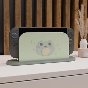 Puede incluir: Una consola Nintendo Switch gris con un soporte verde claro con un diseño de rana de dibujos animados. El soporte está sobre una superficie blanca, con un fondo de listones de madera. La consola tiene botones azules y rojos.
