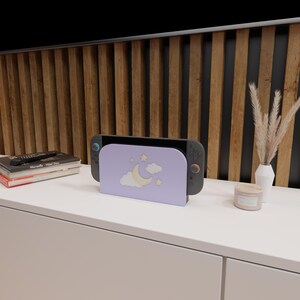 May include: A lavender-colored gaming console stand featuring a crescent moon, stars, and cloud design. The stand holds a black and gray gaming console. The stand is on a white surface, with a wooden slatted wall in the background.