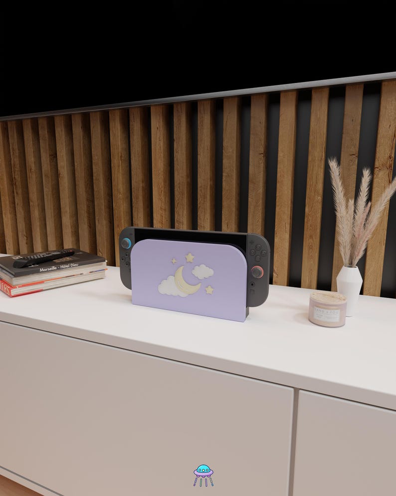 May include: A lavender-colored gaming console stand featuring a crescent moon, stars, and cloud design. The stand holds a black and gray gaming console. The stand is on a white surface, with a wooden slatted wall in the background.