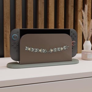 Könnte beinhalten: Eine graue Nintendo Switch-Konsole mit blauen und roten Controllern wird in einem braunen Ständer präsentiert. Der Ständer hat ein florales Design in Weiß und Grün. Der Ständer steht auf einer hellgrauen Oberfläche mit Holzgrund.